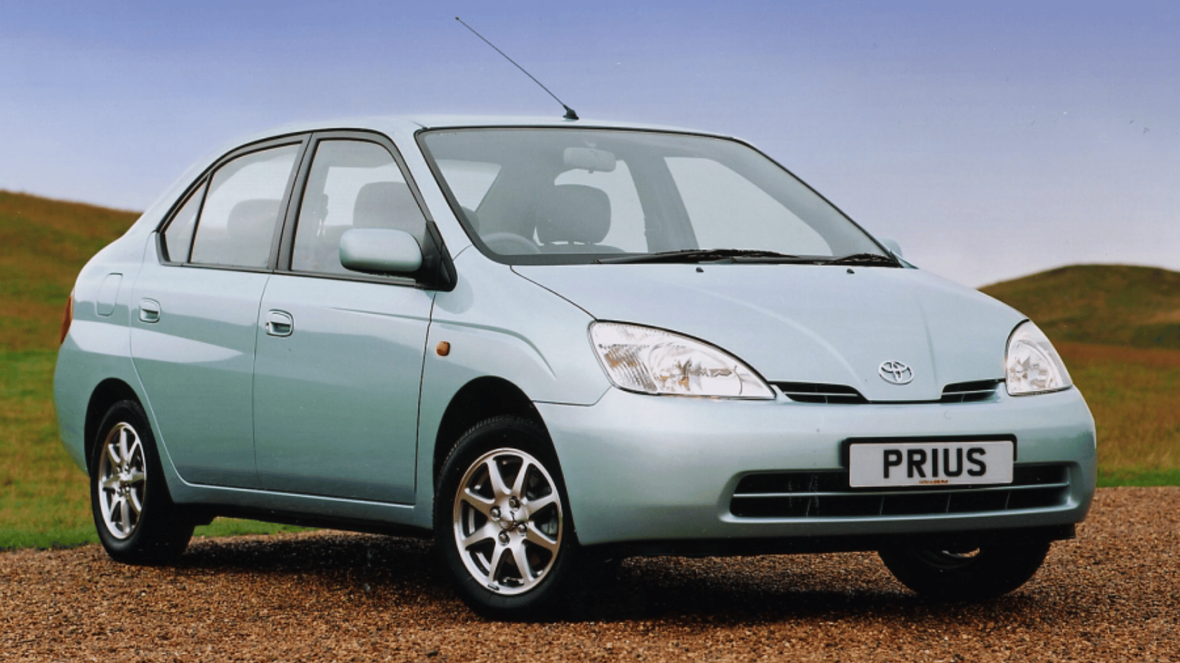 Light blue Toyota Prius hybrid car parked on gravel road with grassy hills and clear sky in background.