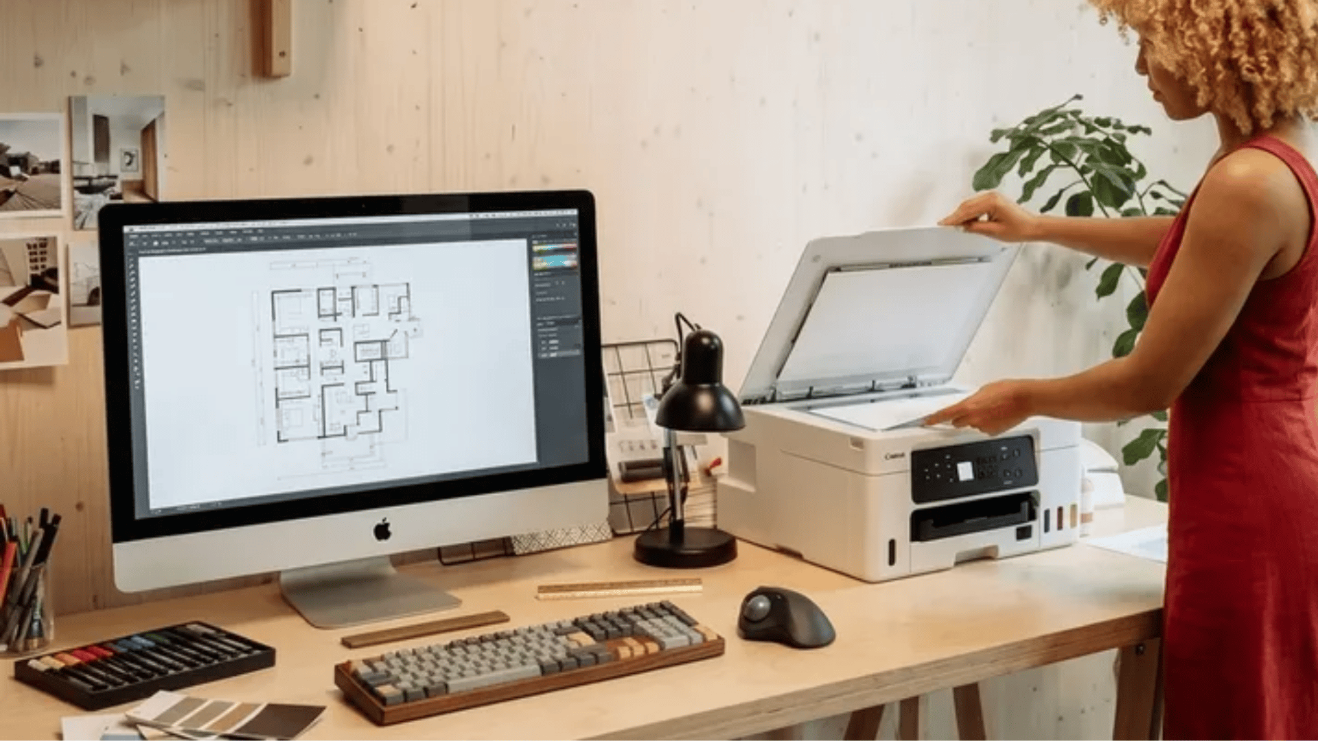 Person using a desktop scanner printer on a wooden desk beside a computer showing a floor plan in a home office setup, illustrating how to scan from printer to computer.