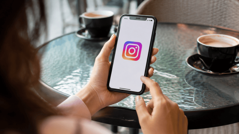 Hand holding smartphone showing Instagram logo on screen in a café setting with coffee cups on table
