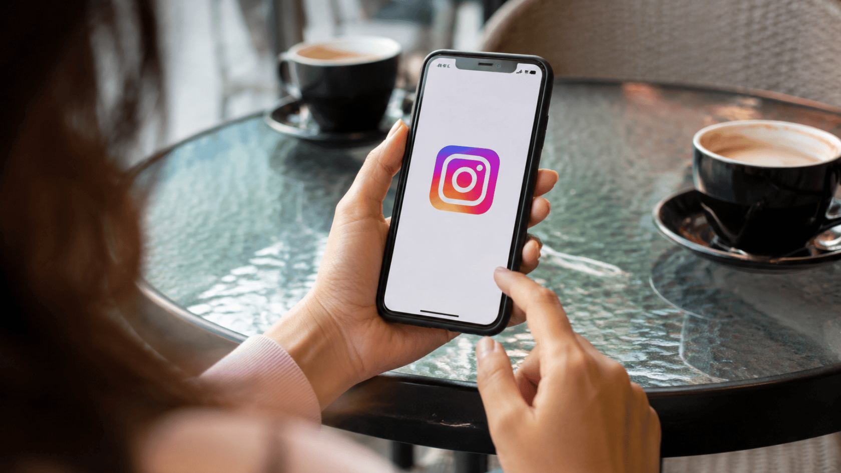 Hand holding smartphone showing Instagram logo on screen in a café setting with coffee cups on table