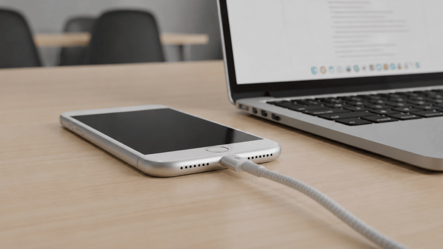 iPhone with home button plugged into a charging cable on a wooden desk next to a laptop.