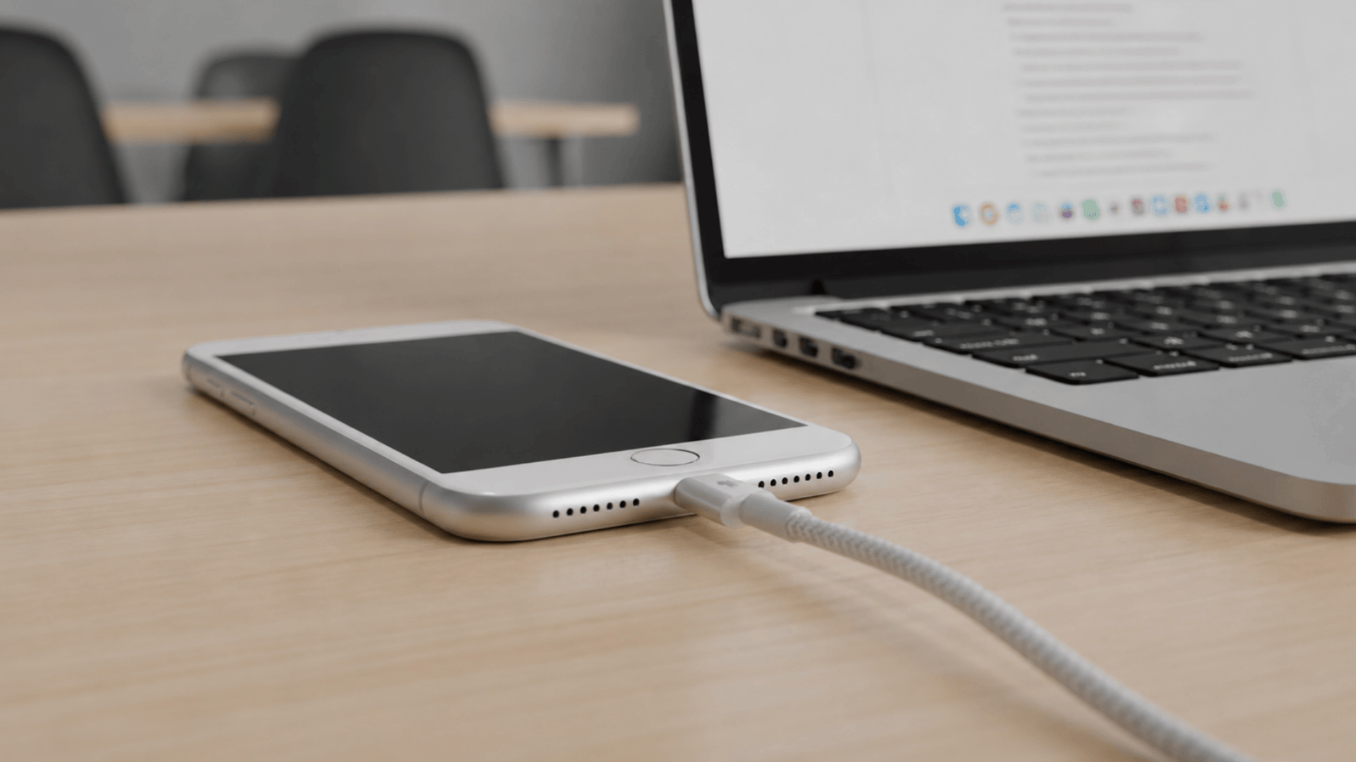 iPhone with home button plugged into a charging cable on a wooden desk next to a laptop.