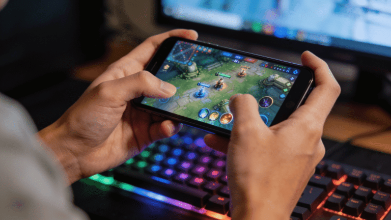 Hands holding a smartphone while playing a mobile game, with a blurred RGB-lit gaming setup in the background