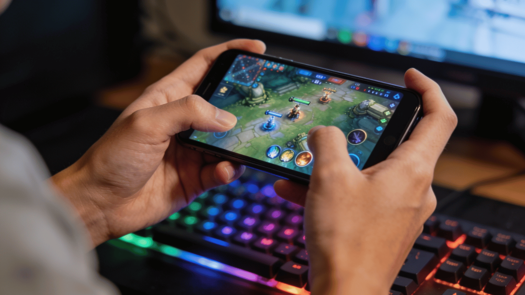 Hands holding a smartphone while playing a mobile game, with a blurred RGB-lit gaming setup in the background