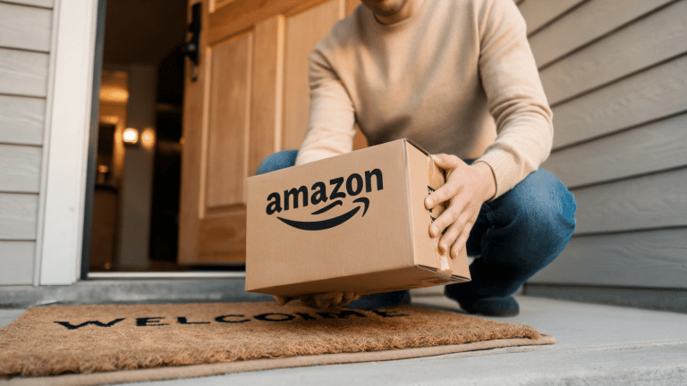 A person kneeling at a home entrance holding a cardboard Amazon delivery package on a doormat near an open front door.