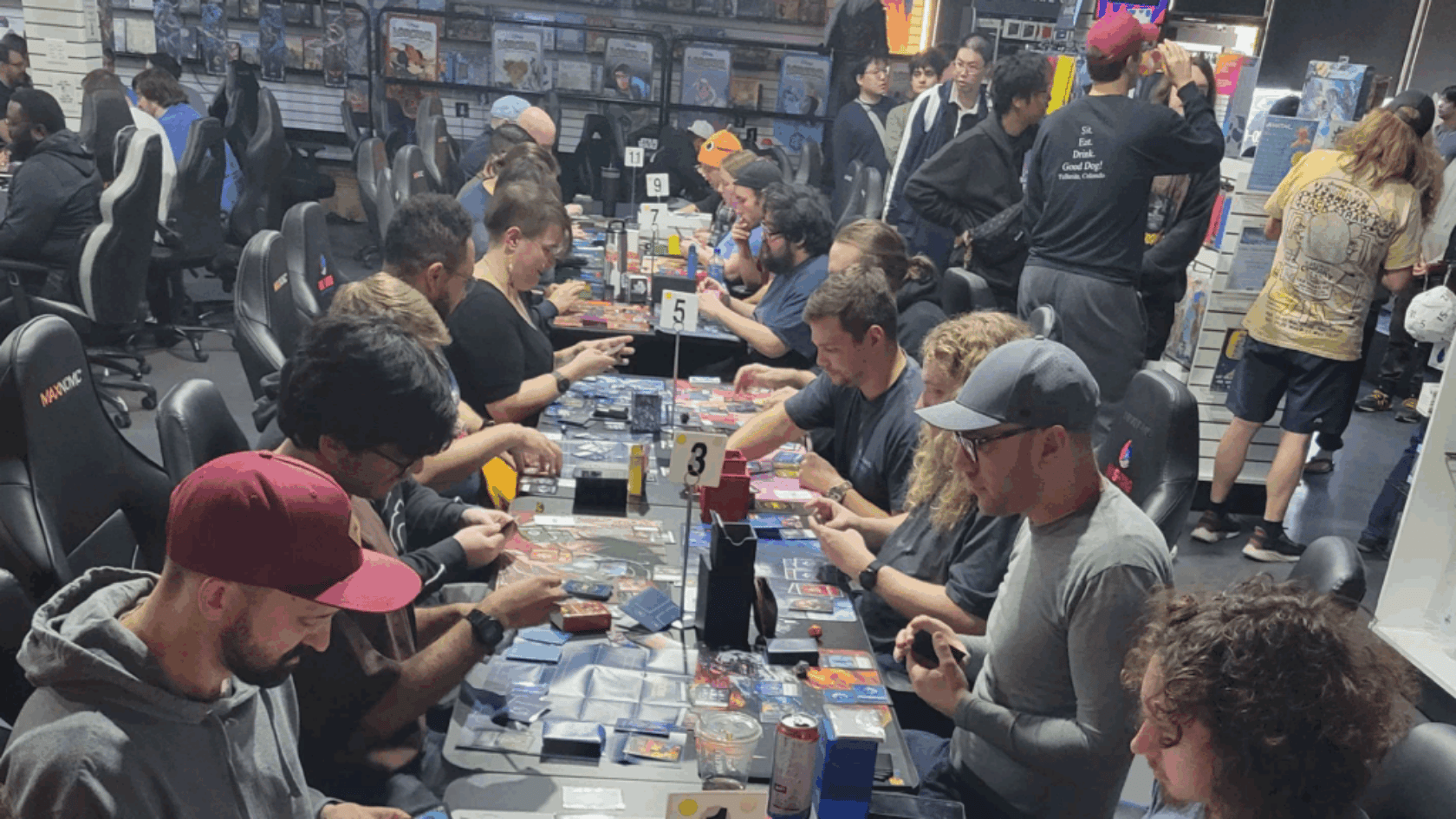 People seated at long tables in a gaming store playing trading card games with cards deck boxes and numbered table signs