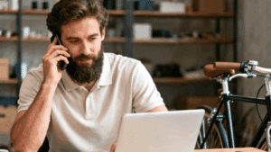 Bearded man in a white polo shirt talking on the phone while working on a laptop, with a bicycle parked in the background