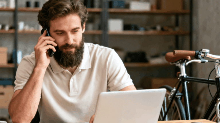 Bearded man in a white polo shirt talking on the phone while working on a laptop, with a bicycle parked in the background