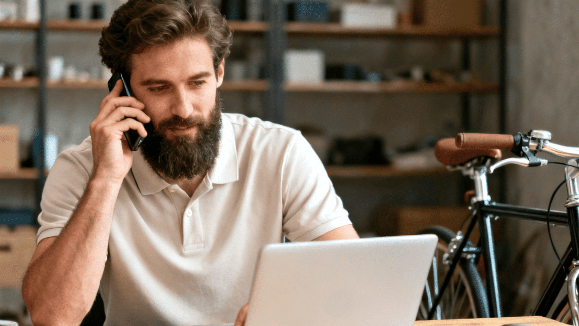 Bearded man in a white polo shirt talking on the phone while working on a laptop, with a bicycle parked in the background