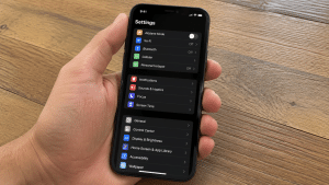 Hand holding iPhone showing Settings app screen in dark mode over wooden table