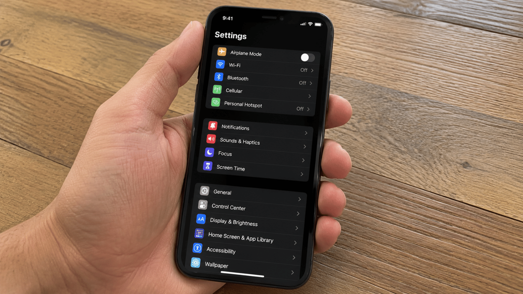 Hand holding iPhone showing Settings app screen in dark mode over wooden table