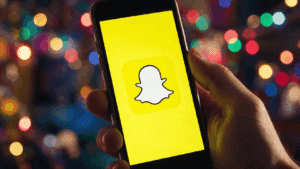Hand holding a smartphone displaying the Snapchat logo against a blurred colorful background.