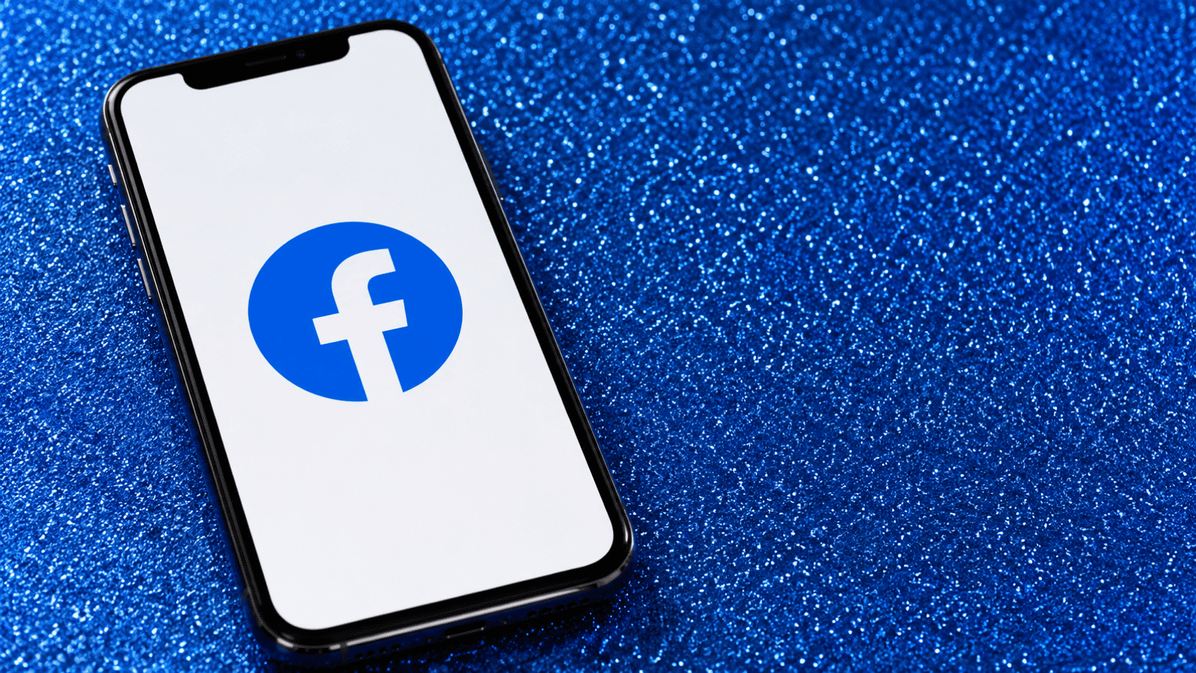 Smartphone showing Facebook logo on screen placed on blue glitter background with diagonal phone orientation front view
