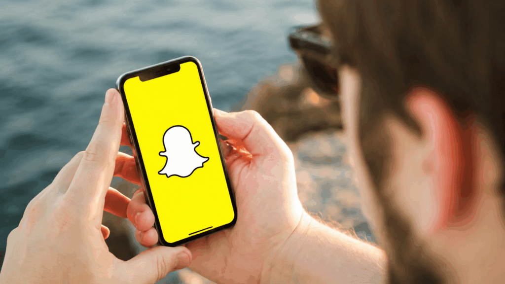 Person holding a smartphone displaying the Snapchat logo near a waterfront background.