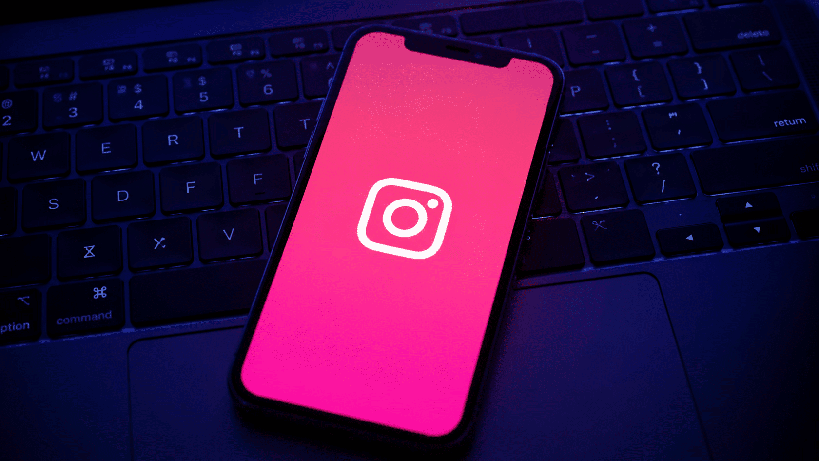 Smartphone displaying Instagram logo on a dark keyboard with pink screen glow and blue ambient lighting.