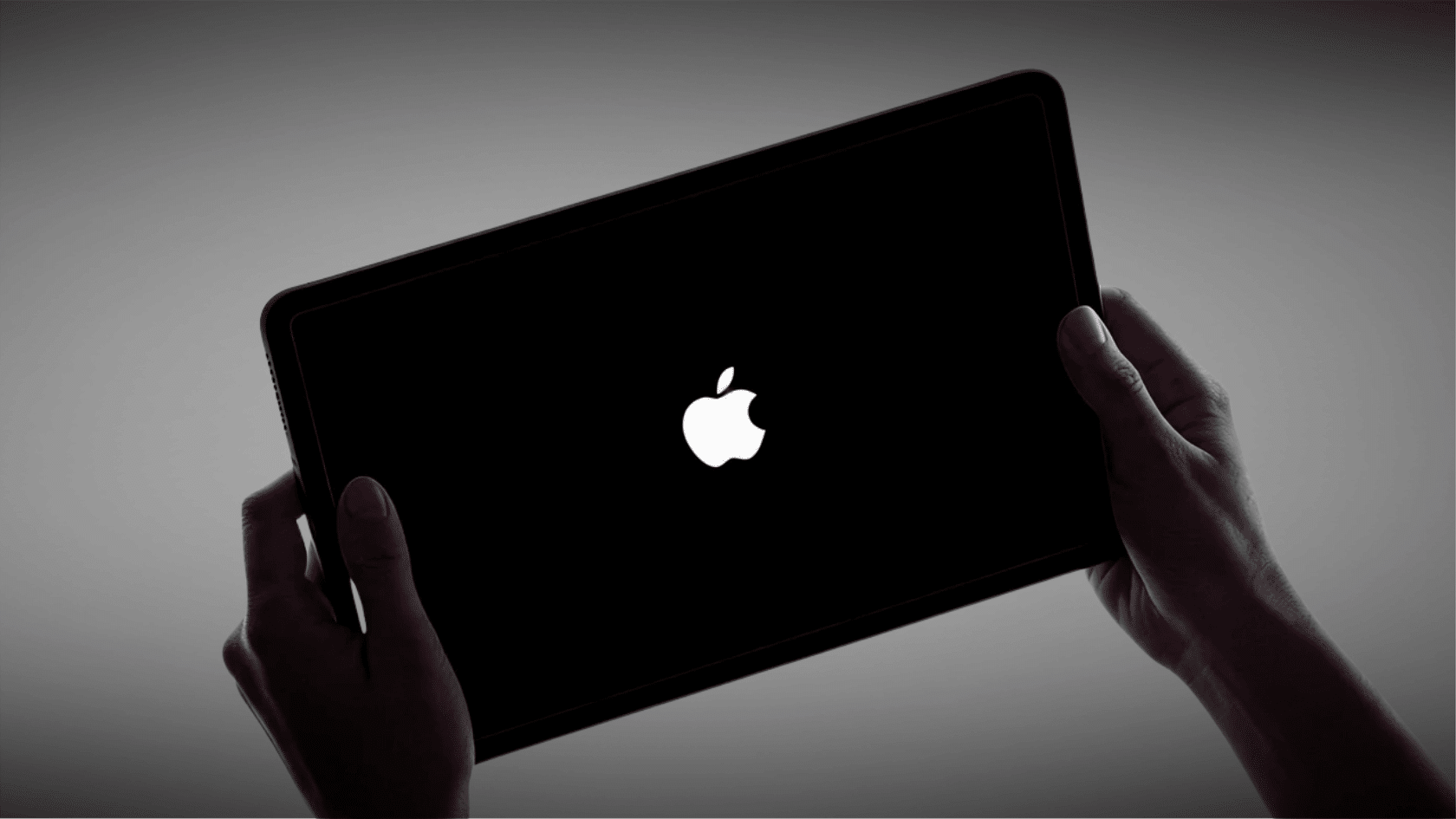 Hands holding an iPad with a black screen displaying the Apple logo during startup.