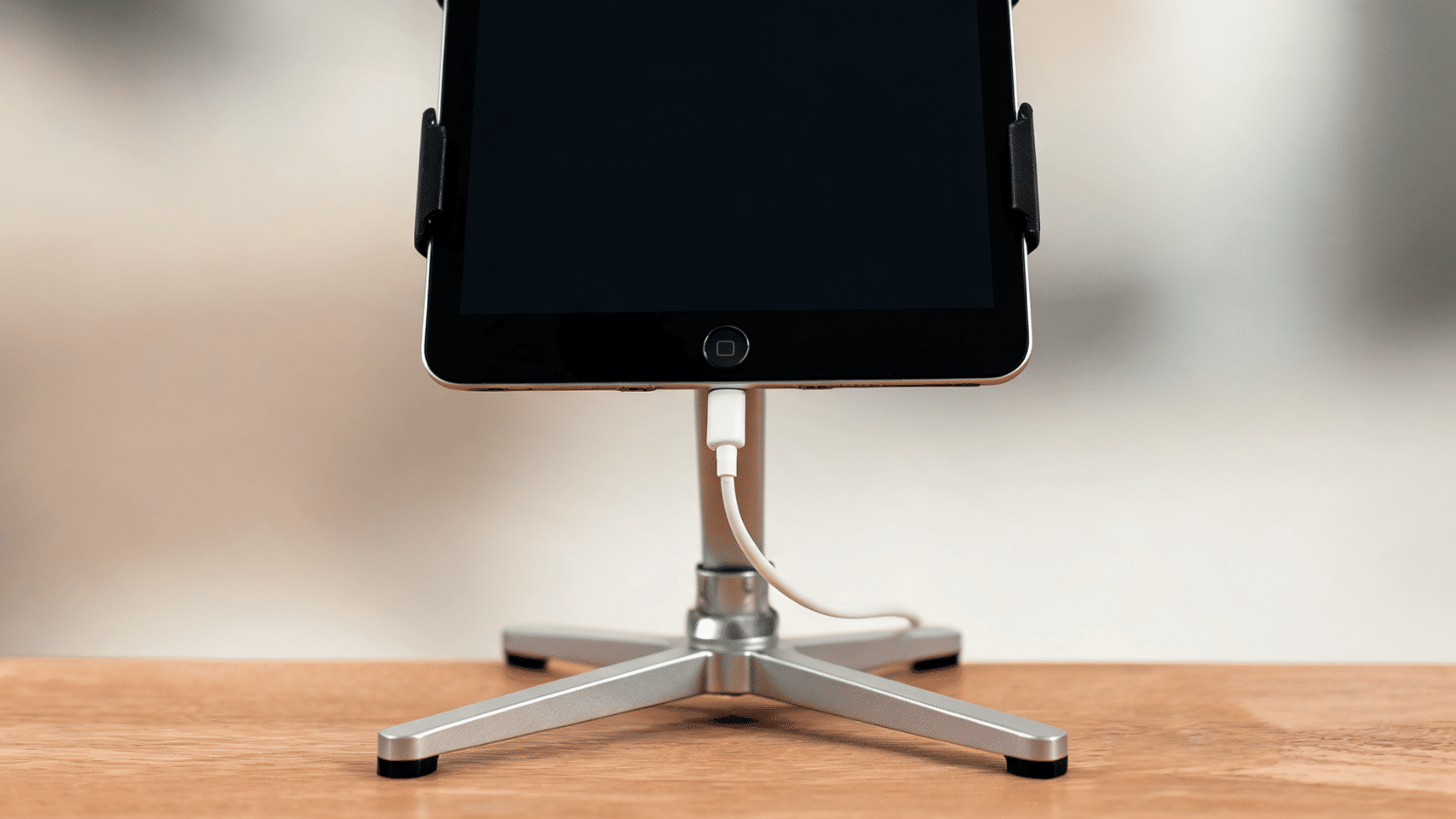 iPad charging on a stand with a cable connected on a wooden desk surface.